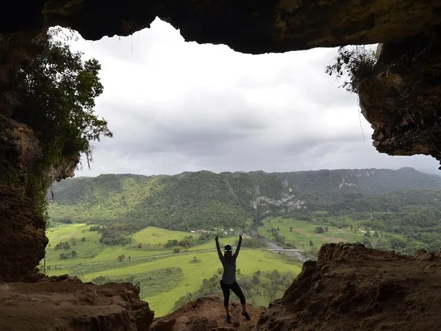 Woman standing in a cave entrance with arms raised facing a valley.